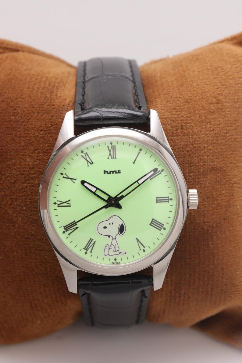 May include: A silver-toned wristwatch with a light green face featuring Snoopy. The watch has black hands and Roman numeral hour markers. The black leather band complements the watch face. The word "INDIA" is printed on the watch face.