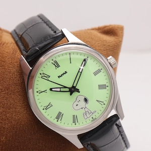 May include: A silver-toned wristwatch with a light green face featuring Roman numerals and a Snoopy graphic. The watch has black hands and a black leather band. The watch is set on a brown surface.