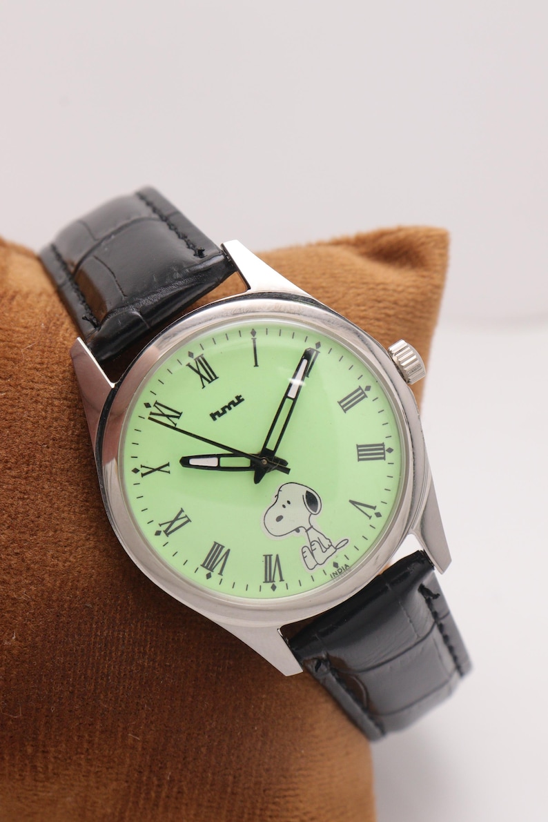 May include: A silver-toned wristwatch with a light green face featuring Roman numerals and a Snoopy graphic. The watch has black hands and a black leather band. The watch is set on a brown surface.