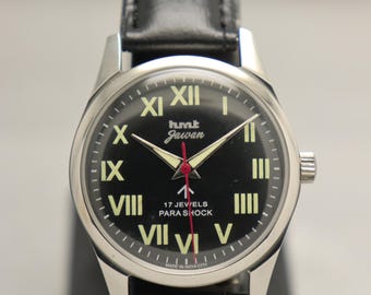 Puede incluir: Un reloj de pulsera HMT Jawan vintage con esfera negra y caja plateada. El reloj presenta agujas e indicadores horarios luminosos, una correa de cuero negro y el texto "hmt Jawan" en la esfera. También tiene el texto "17 JEWELS PARA SHOCK".
