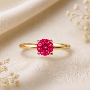 May include: A gold ring featuring a round, vibrant pink gemstone set in a four-prong setting. The band is smooth and polished, showcasing a minimalist design that highlights the brilliant, saturated color of the central stone in a classic style.