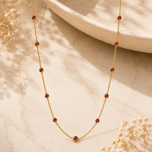 Puede incluir: Un collar de cadena dorada con piedras preciosas rojas redondas, engastadas en bisel y espaciadas uniformemente. La delicada cadena de metal conecta cada engaste circular, creando un patrón constante de oro y rojo intenso a lo largo de la pieza.