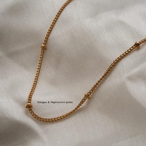 May include: A gold-colored necklace with a delicate chain design. The necklace features small, decorative knots at intervals along the chain. The text "Unique & Impressive Gems" is visible in the image.