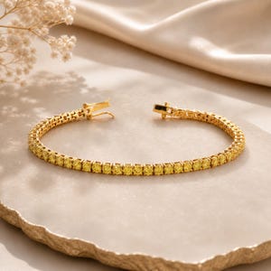 Könnte beinhalten: Ein goldfarbenes Tennisarmband mit einer durchgehenden Reihe kleiner, runder, leuchtend gelber Steine in einer Metallfassung. Das Armband verfügt über einen sicheren Kastenschloss-Verschluss am Ende, der in geöffneter Position dargestellt ist.