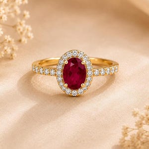 May include: A gold ring featuring a central oval-cut deep red gemstone surrounded by a halo of small round white stones. The band is also set with a row of small round white stones along each side, creating a bright and reflective appearance.