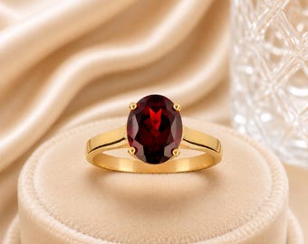 May include: A gold ring featuring a large, oval-cut, deep red garnet gemstone held in a four-prong setting. The band is smooth and polished, showcasing a classic design that highlights the vibrant color and clarity of the central stone.