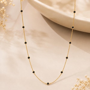 Könnte beinhalten: Eine zarte Goldkettenhalskette mit gleichmäßig verteilten, kleinen, runden, schwarzen Steinen, die in Goldfassungen eingefasst sind. Die Kette hat eine feine Gliederkonstruktion und zeigt eine Reihe dunkler, kreisförmiger Akzente entlang der gesamten Länge.