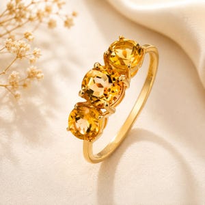 Puede incluir: Un anillo de oro con tres piedras preciosas de citrino amarillo de corte redondo dispuestas en fila. Las piedras están montadas en un engaste de garras sobre una banda de oro fina y pulida. El diseño resalta las superficies facetadas de las piedras.