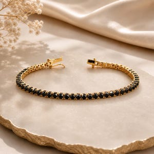 Op de afbeelding: Een goudkleurige tennisarmband met een doorlopende rij ronde, gefacetteerde zwarte stenen in een metalen zetting. De armband is voorzien van een veilige baksluiting met een veiligheidsklepje en heeft een strak en uniform ontwerp.