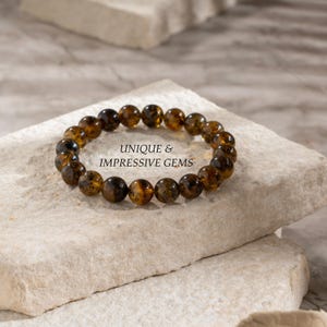 May include: A bracelet made of amber-coloured, spherical gemstones. The bracelet is presented on a light-coloured, textured surface. The image includes the text "UNIQUE & IMPRESSIVE GEMS".