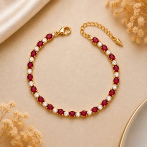 May include: A gold-tone tennis bracelet featuring alternating oval-cut deep red stones and round clear stones. The bracelet includes a lobster claw clasp and an adjustable chain extension with a small pointed charm at the end of the chain.
