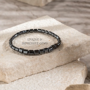 May include: A dark grey beaded bracelet with square beads sits on a light-coloured stone surface. The bracelet is made of small, evenly sized beads. The text "UNIQUE & IMPRESSIVE GEMS" is visible in the image.