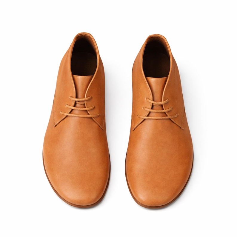 Puede incluir: Un par de botines chukka de cuero color marr&oacute;n claro, vistos desde arriba, sobre un fondo blanco. Los botines tienen un dise&ntilde;o cl&aacute;sico con cierre de cordones y punta redondeada. El cuero tiene una textura suave y un color c&aacute;lido y natural.