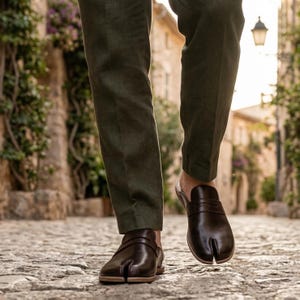 Pode incluir: Sapatos de couro castanho escuro com biqueira dividida e uma tira. Os sapatos têm um design único com um compartimento separado para o dedo grande. Usados com calças verde-escuras, numa rua de calçada.