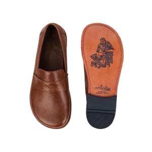 May include: Brown leather loafers with a classic design, featuring a rounded toe and a decorative strap across the vamp. The sole of one shoe is visible, displaying a craftsman illustration and the brand name.