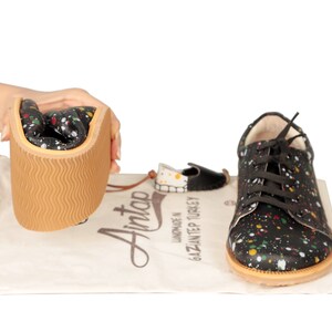 Op de afbeelding: Zwarte leren sneakers met een kleurrijk verfspattenpatroon. De schoenen hebben een bruine rubberen zool en witte veters. De schoenen worden getoond met een witte trekkoordtas met de tekst "Handgemaakt in Gaziantep Turkije" en het logo "Aintao".