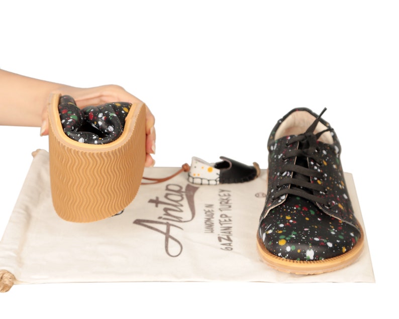 Op de afbeelding: Zwarte leren sneakers met een kleurrijk verfspattenpatroon. De schoenen hebben een bruine rubberen zool en witte veters. De schoenen worden getoond met een witte trekkoordtas met de tekst "Handgemaakt in Gaziantep Turkije" en het logo "Aintao".