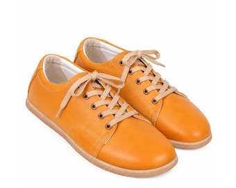 May include: A pair of mustard yellow low-top sneakers featuring a smooth finish, rounded toes, and light beige laces threaded through metal eyelets. The shoes have a white interior lining and a flat, light-colored rubber sole for a clean look.