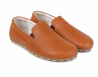 May include: A pair of tan leather slip-on shoes featuring a smooth, rounded toe design and a visible white stitched detail along the edge of the sole. The interior lining is white, and the shoes have a low-profile silhouette with no laces.