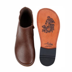 May include: A brown leather ankle boot with a side zipper and a smooth rounded toe. Beside it, the sole of the boot is visible, featuring a stamped illustration of a shoemaker, the text Aintap, and the words Handmade in Gaziantep-Türkiye.