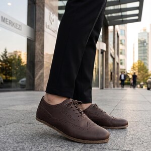 May include: A pair of dark brown leather oxford shoes featuring classic wingtip brogue detailing, thin matching laces, and a low-profile sole. The shoes are worn with slim-fit black trousers, showcasing a structured design with visible stitching.