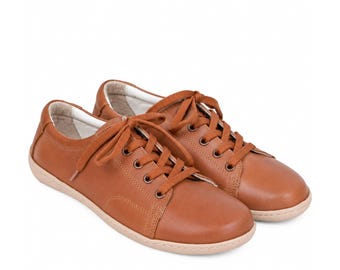 May include: A pair of low-top sneakers in a solid tan brown color. These shoes feature a smooth leather-like exterior, matching tan laces threaded through metal eyelets, a rounded toe box, and a light beige rubber sole with a textured edge.