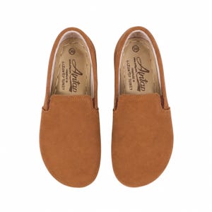 May include: A pair of tan slip-on shoes with a rounded toe. The interior insole features the text Aintap, size 39, and Handmade in Gaziantep Turkey. The shoes have a smooth, solid-colored exterior and a light-colored interior lining with visible stitching.