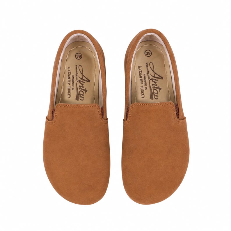 May include: A pair of tan slip-on shoes with a rounded toe. The interior insole features the text Aintap, size 39, and Handmade in Gaziantep Turkey. The shoes have a smooth, solid-colored exterior and a light-colored interior lining with visible stitching.