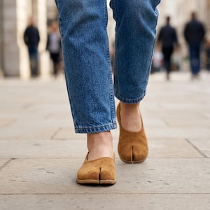 Può includere: Una persona indossa jeans blu e scarpe marroni con punta divisa. Le scarpe hanno un tacco basso e un design unico che separa l'alluce. Le scarpe sono realizzate in un materiale simile alla pelle scamosciata.