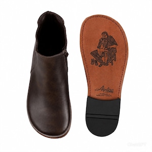 Puede incluir: Un botín marrón oscuro con cremallera lateral y punta redondeada. Junto a él se encuentra la suela del zapato, con una superficie de cuero color canela, un tacón negro, una ilustración estampada de un zapatero y el texto Aintab Handmade in Gaziantep Turkiye.