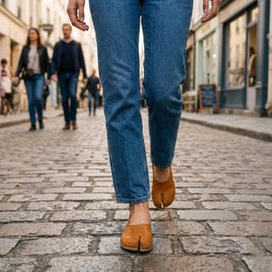 May include: A person wearing blue jeans and brown split-toe shoes walks on a cobblestone street. The shoes have a unique design with a separate space for the big toe. Other people are walking in the background.