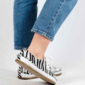 May include: A pair of black and white zebra print low-top sneakers with white laces. The shoes are worn by a person wearing blue jeans.