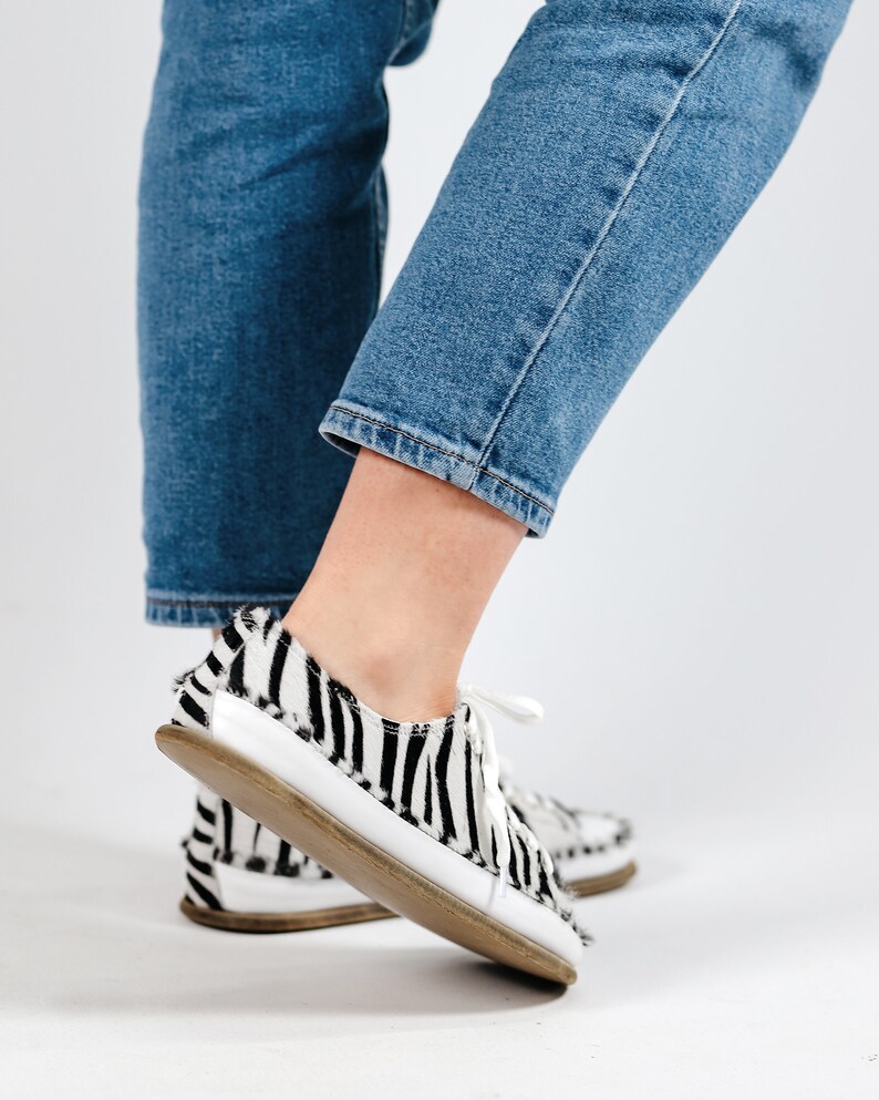 May include: A pair of black and white zebra print low-top sneakers with white laces. The shoes are worn by a person wearing blue jeans.