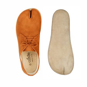 May include: Orange suede split-toe shoe with laces, next to a matching insole. The shoe has a unique design with a separate space for the big toe. The insole is beige. The shoe has the brand name "Liz Baa" printed on the insole.