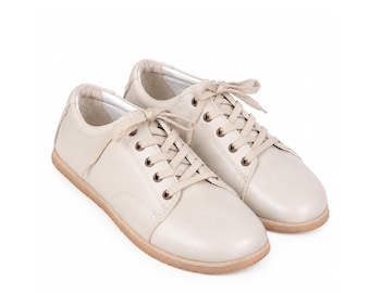May include: A pair of low-top sneakers in a solid off-white color. These shoes feature a smooth upper, matching off-white laces threaded through metal eyelets, a rounded toe, and a flat, light tan rubber sole with a textured edge for traction.
