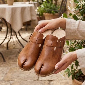 May include: A pair of brown leather clogs with a split-toe design. The clogs have a buckle closure and are held by a person. The clogs are a warm brown color and appear to be new.