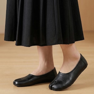 May include: Black leather split-toe ballet flats. The shoes have a rounded toe and a low heel. The design features a split between the big toe and the other toes. The flats are worn with a black skirt.