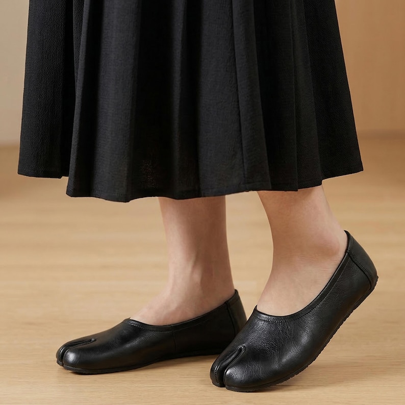 May include: Black leather split-toe ballet flats. The shoes have a rounded toe and a low heel. The design features a split between the big toe and the other toes. The flats are worn with a black skirt.