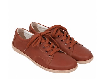 May include: A pair of low-top sneakers in a solid rust-brown color. These shoes feature matching brown laces, metal eyelets, a rounded toe, and a light beige flat sole. The interior lining of the sneakers is a light cream color.