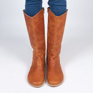 May include: A pair of knee-high brown leather boots with a light tan sole. The boots have a zip at the back and are worn with blue jeans. The boots have a classic design and are made of leather.