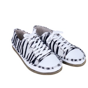 May include: A pair of zebra-striped sneakers with white laces and toe caps. The black and white pattern covers the sides and heel of the shoes. The soles are a light brown color.