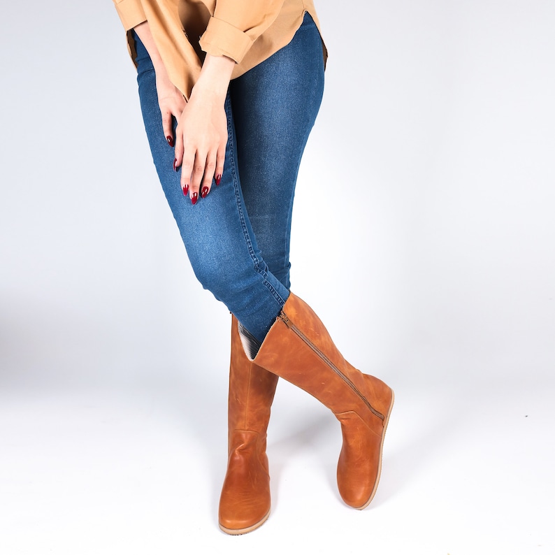 May include: A pair of tall, brown leather boots with a side zip. The boots are worn with blue jeans and a tan shirt. The boots appear to be made of a soft leather material and have a flat sole.
