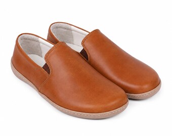 May include: A pair of tan slip-on shoes featuring a smooth leather-like exterior, a rounded toe design, and a light-colored interior lining. The shoes have a low-profile silhouette with visible stitching along the sole and side panels.