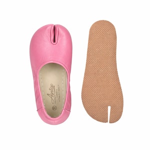 May include: A pair of pink split-toe ballet flats with a separate brown sole. The pink leather shoe has a rounded toe and a split between the big toe and the other toes. The sole is a separate piece with a textured surface.