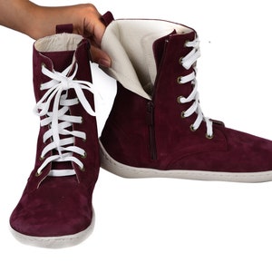 May include: Burgundy suede ankle boots with white laces and a zipper on the side. The boots have a white rubber sole.