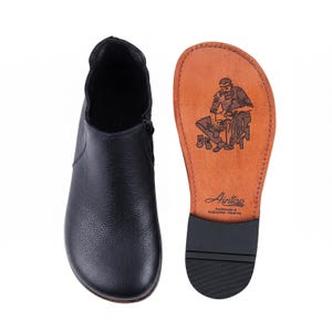 May include: A black leather ankle boot with a textured surface and a side zipper, shown alongside its tan leather sole. The sole features a stamped illustration of a shoemaker and the text Aintap, Handmade in Gaziantep-Türkiye.
