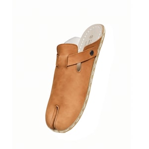 May include: A tan leather clog-style shoe with a split toe design. The shoe features a strap across the top secured with a dark button. The sole is stitched with white thread. The interior is white.