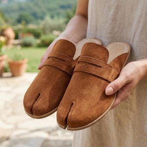 May include: A pair of brown suede split-toe sandals. The sandals have a strap across the top with a decorative edge. The soles are light-colored with white stitching. The sandals are held in front of a blurred outdoor background.