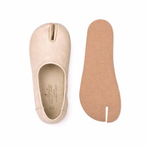 May include: Tan leather split-toe shoes, with a separate sole. The shoe has a rounded toe with a split for the big toe. The sole is a light brown color with a textured surface. The shoes are designed for comfort and flexibility.