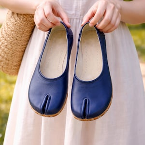 May include: A pair of navy blue split-toe shoes. The shoes have a rounded toe design with a split between the big toe and the other toes. The shoes are held up by a person. The shoes are made of a smooth material.